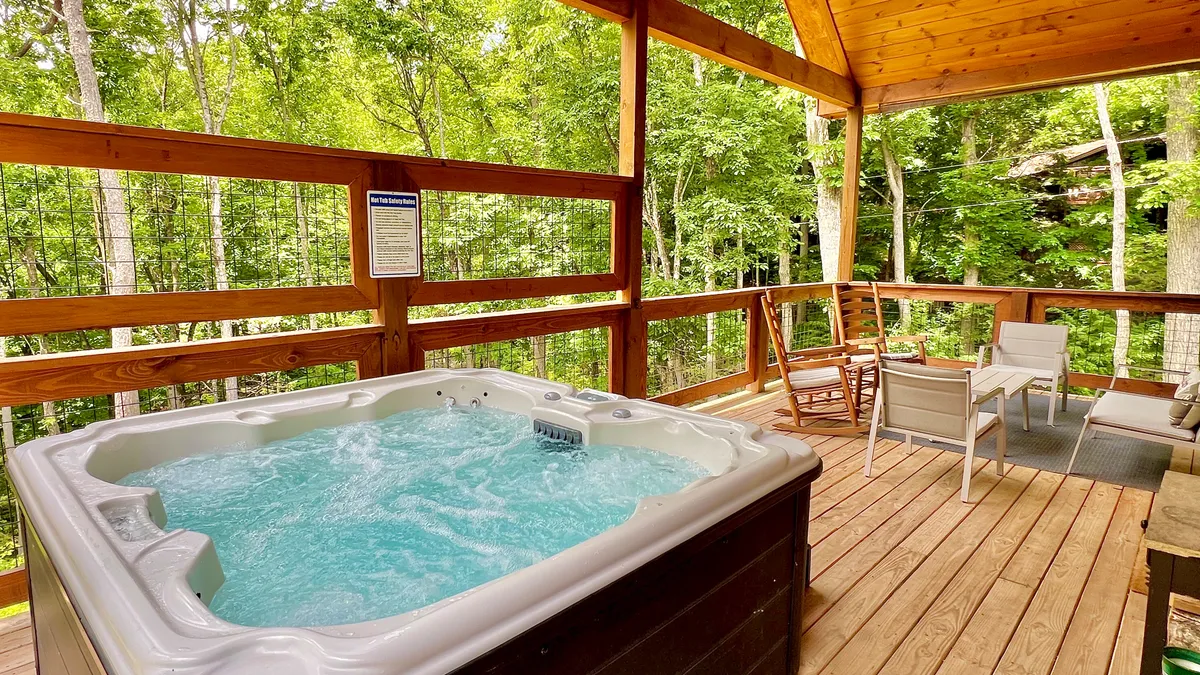 The image showcases a classic wooden cabin nestled among lush trees, with a spacious deck featuring a bubbling hot tub, comfortable seating area, and sweeping views of the serene Smoky Mountain hills at sunset.