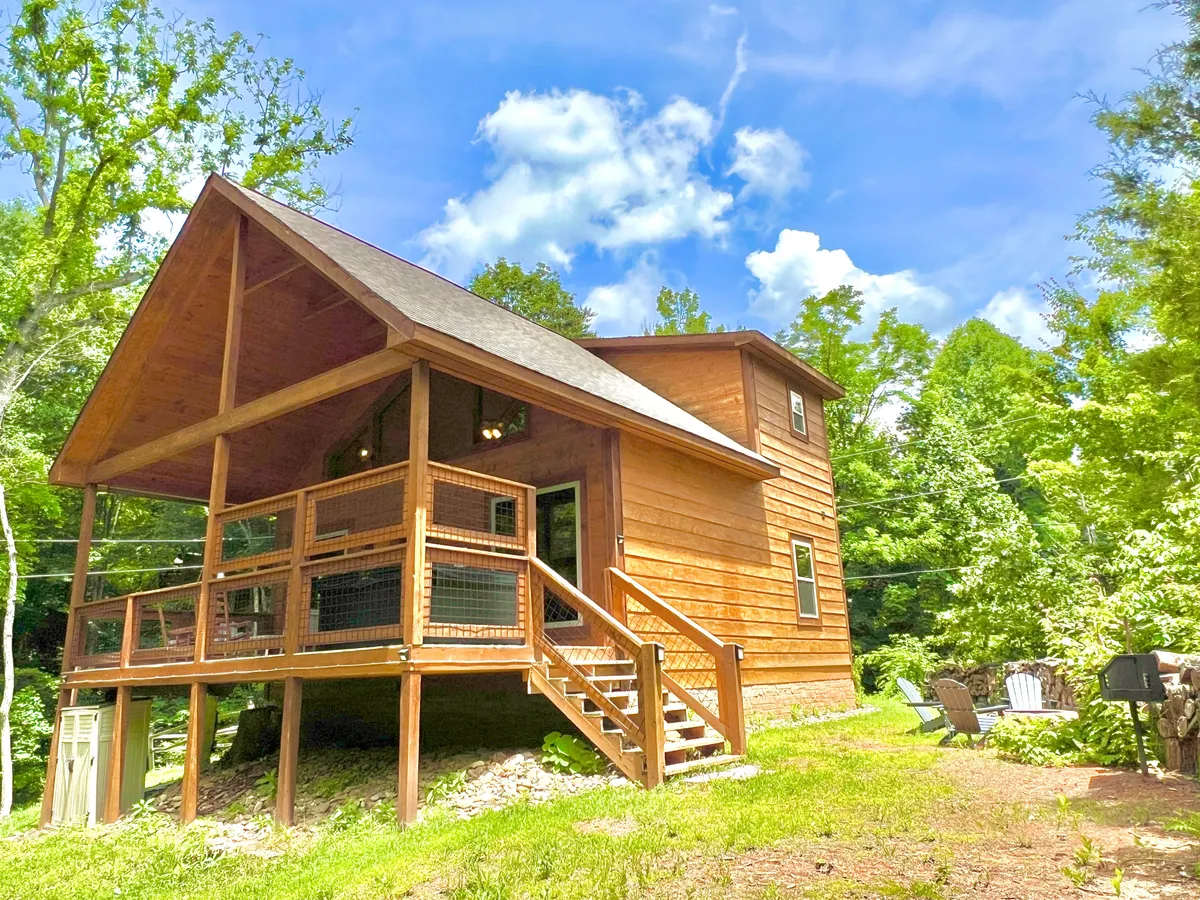 A wide-angle view of the wooden cabin's exterior and its spacious deck, framed by peaceful woodland scenery. The deck includes a welcoming outdoor seating area, perfect for relaxation or gathering, and hints at the inviting atmosphere for barbecues, morning coffee, and stargazing.