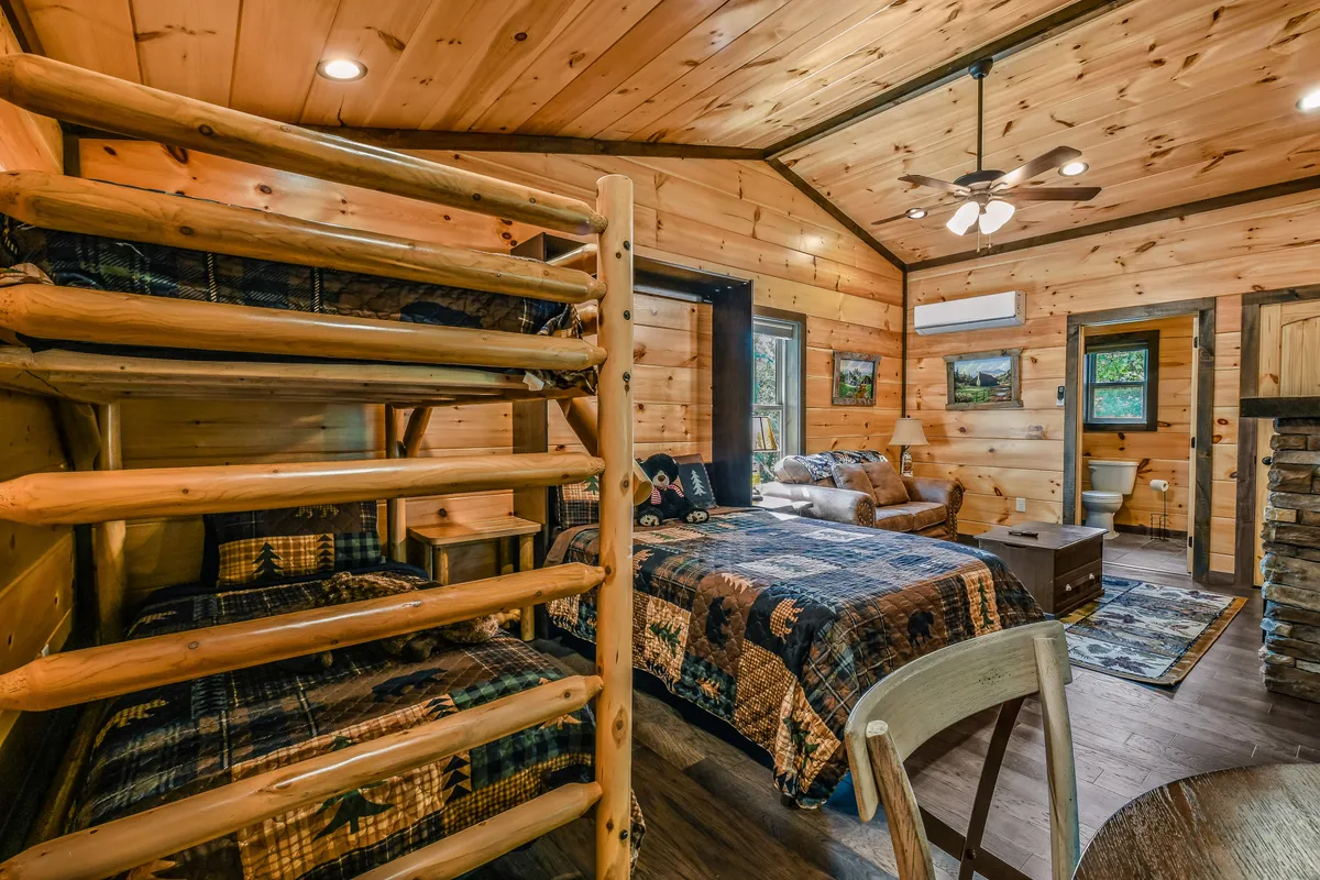 An inviting loft bedroom featuring a queen bed, a twin bunk, and a cozy sitting area with wooden accents. The layout is open and welcoming, ideal for families or friends, with natural light streaming in and comfortable furnishings throughout.