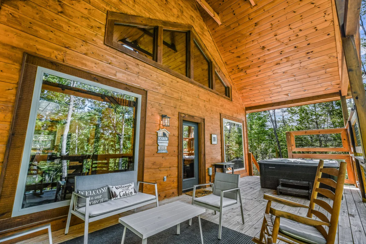 An inviting exterior view of Honeysuckle Hill, featuring a wooden cabin structure with large windows that open onto a spacious deck. The deck is equipped with an outdoor seating area and a hot tub, surrounded by the tranquil greenery of the Smoky Mountains.