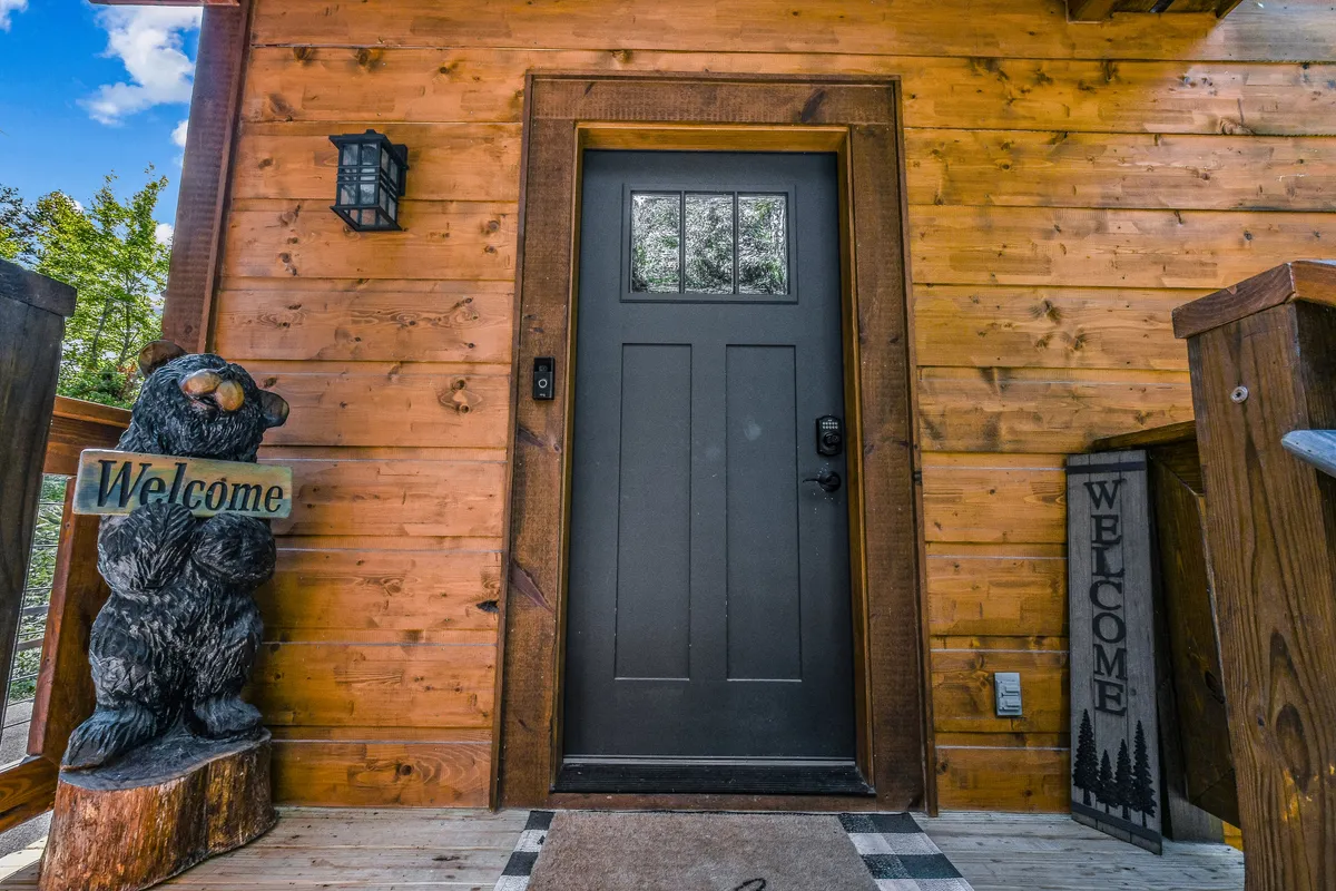This image captures the exterior entrance of the cabin, featuring a dark front door, a bear statue, and a welcoming sign that sets the tone for a homey stay. The porch area invites guests to relax and hints at the natural surroundings of the Smoky Mountains.