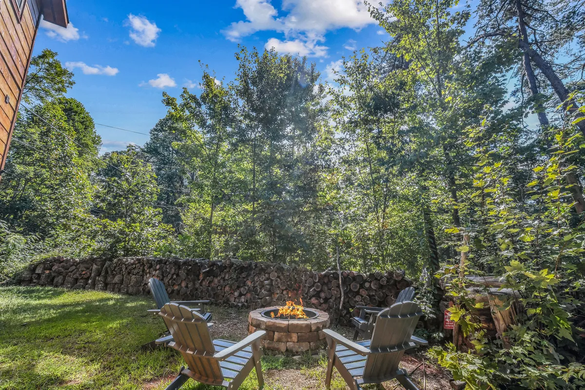 This exterior image features a cozy fire pit surrounded by Adirondack chairs, set against a rustic wooden wall. The area is framed by natural greenery, suggesting a relaxing outdoor gathering spot perfect for friends to unwind under the stars.