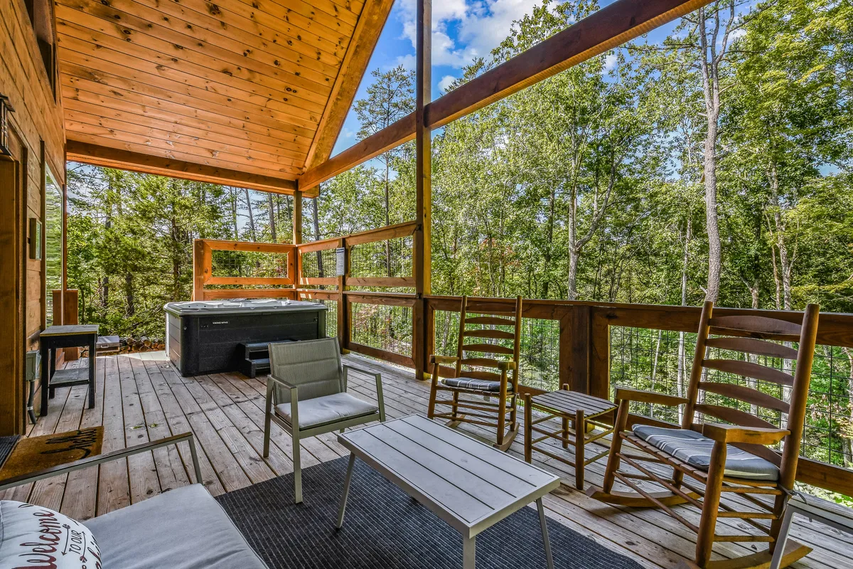 This image showcases a serene wooden deck complete with a bubbling hot tub, classic wooden rocking chairs, and the backdrop of lush greenery. The view captures the tranquil ambiance ideal for unwinding, with seating that invites conversation or relaxation.