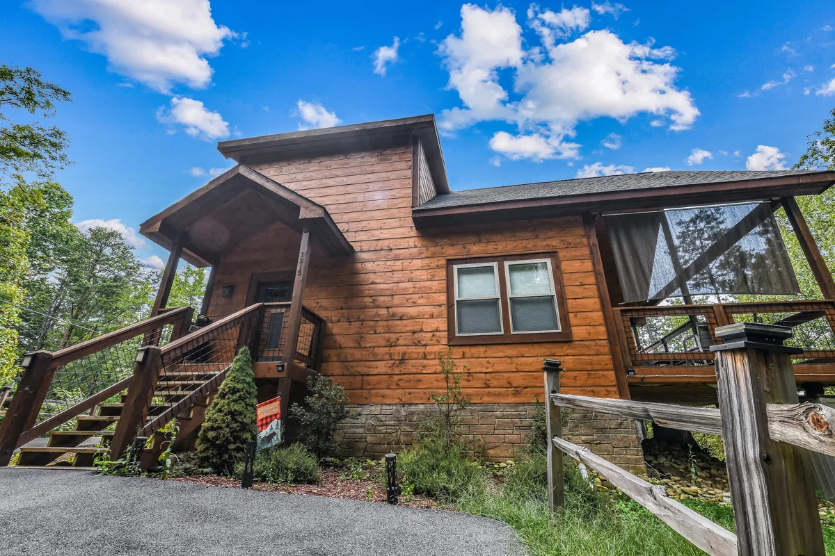 This image displays the welcoming exterior of the cabin, featuring a charming front porch surrounded by lush greenery and set beneath a bright blue sky. The setting hints at a peaceful retreat with plenty of space for gathering outdoors, and the inviting porch suggests a place to relax after a day of adventure.