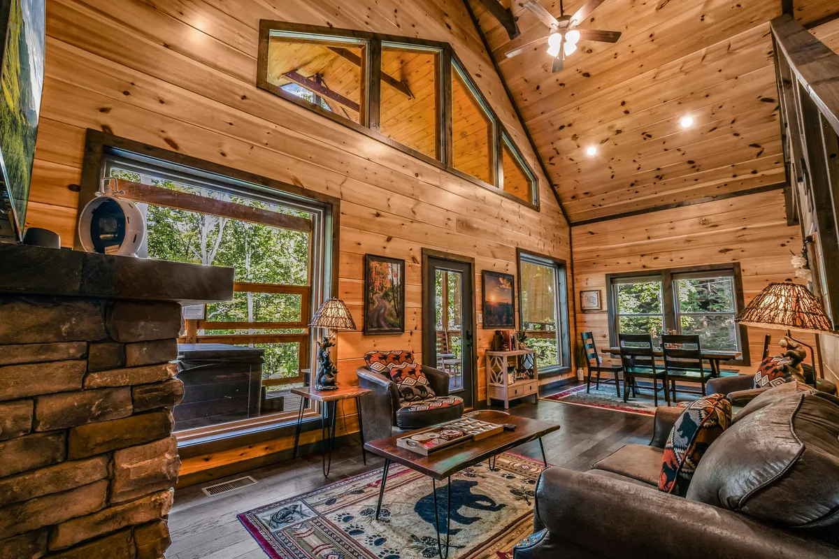 A spacious open-concept interior featuring large windows that allow abundant natural light to fill the living and dining area. The stone fireplace serves as a cozy focal point surrounded by comfortable seating, a welcoming dining table, and decorative rugs, all set against a backdrop of wooded views.
