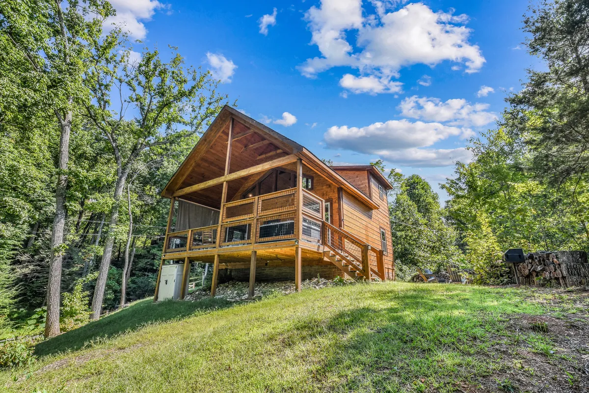 This wide exterior shot captures the wooden cabin structure, deck with railing, and the beautiful surrounding trees under a blue sky. The image conveys tranquility, seclusion, and a sense of arrival—while the deck suggests opportunities for outdoor relaxation and easy living.
