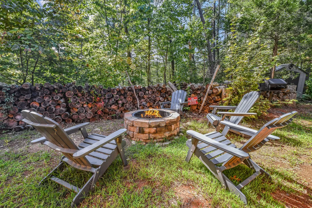 A cozy fire pit area surrounded by adirondack chairs and stacked logs, set among the trees. The scene invites guests to gather for campfire stories, s'mores, and late-night conversations in a warm, inviting setting.